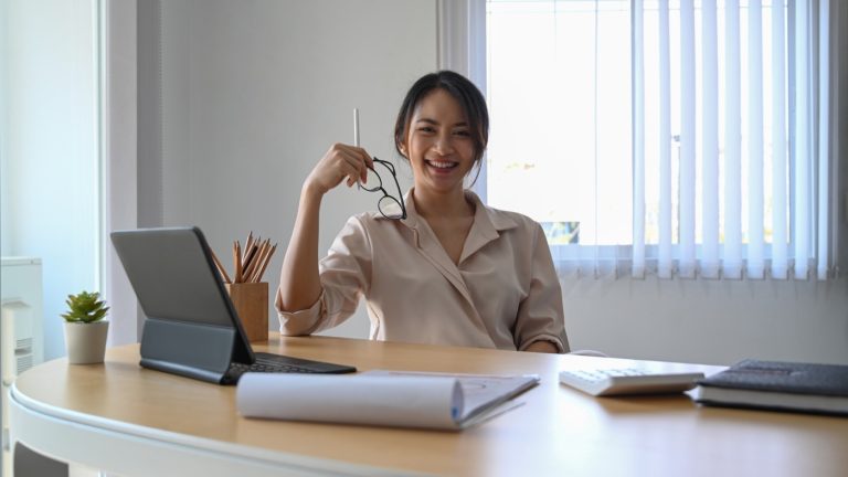 Happy young woman accountant sitting at her workplace. holding her glasses and smiling to camera.