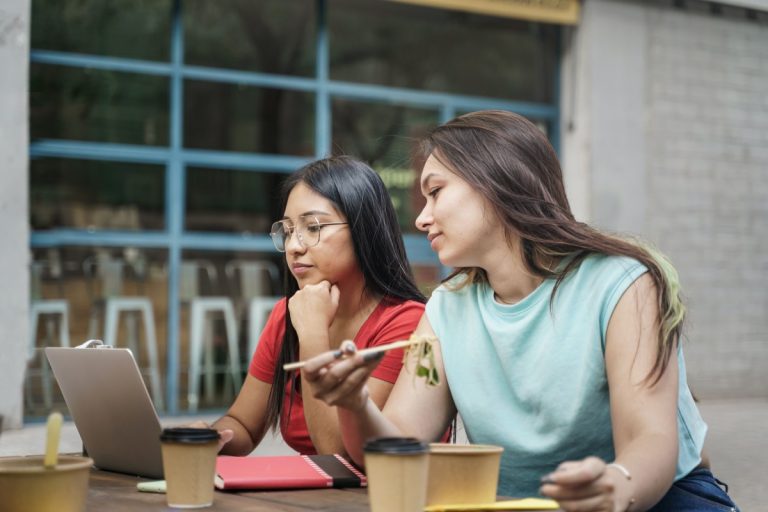 latin woman working on laptop while female colleague eating healthy take out asian noodles